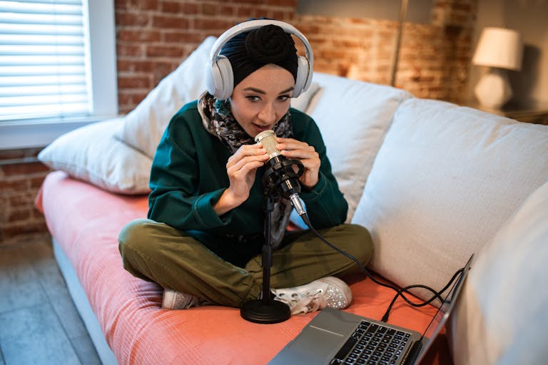 A young woman wearing headphones records a podcast at home using a microphone and laptop.
