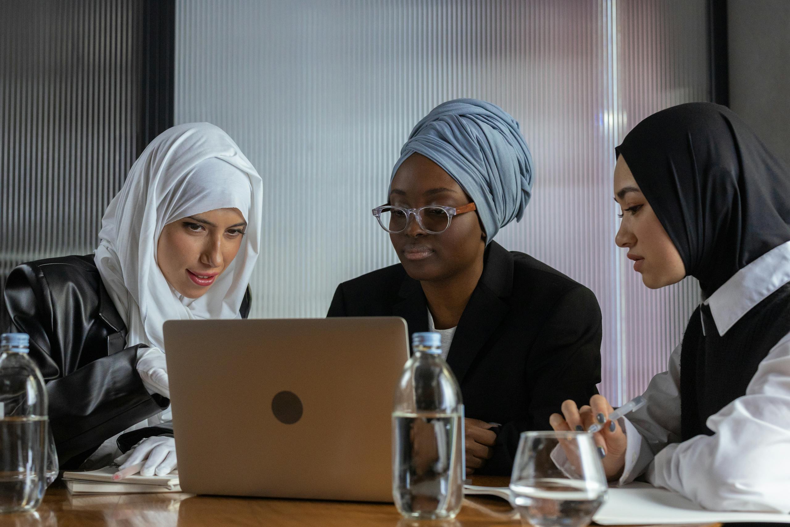 Three women in hijabs and turbans collaborate at a laptop in an indoor office setting.